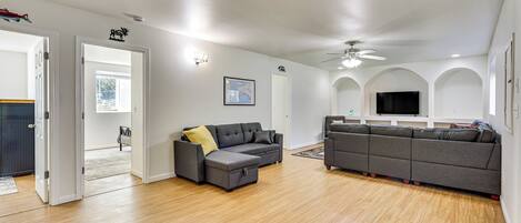 A spacious living area with a grey sectional sofa, a smaller grey sofa, and a wooden floor. There is a ceiling fan, a mounted TV, and a door leading to another room.