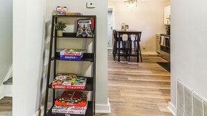 Een hal met een boekenplank vol gezelschapsspellen, een eethoek met tafel en stoelen, en een keuken op de achtergrond.