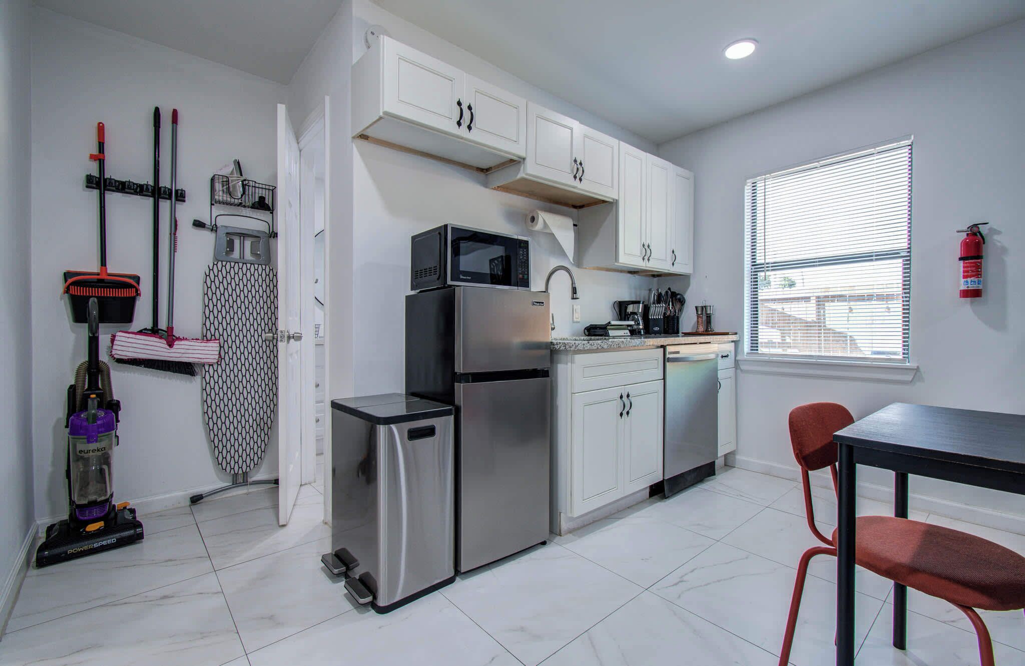 A compact kitchen with white cabinets, a stainless steel refrigerator, a microwave, and a sink. There are two red chairs and a window with blinds.