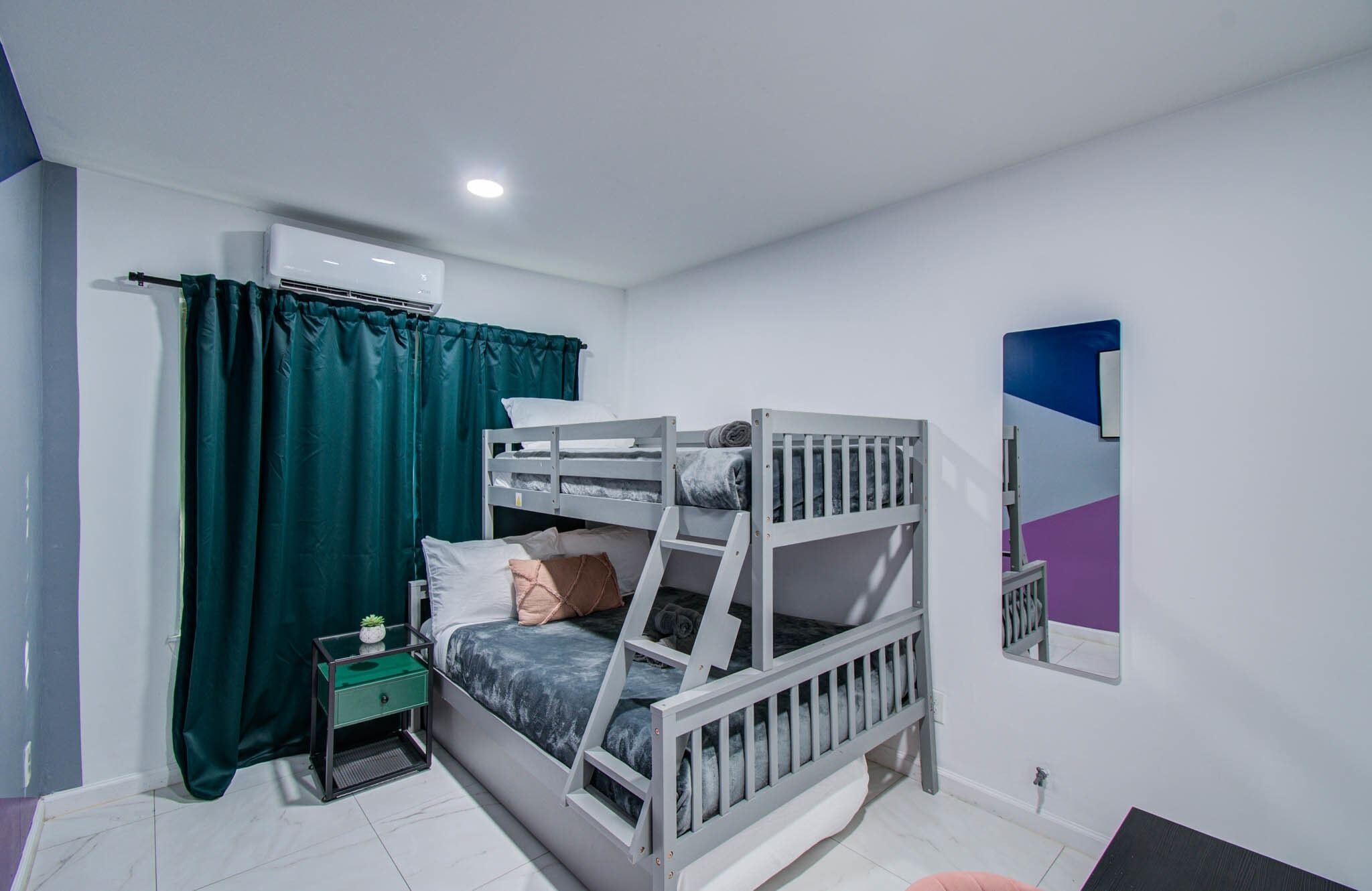 A bunk bed room with a small bedside table, a mirror, and a window with curtains.