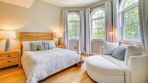 A bedroom with a large bed, a bedside table with lamps, a chair, and a window with curtains.