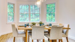 A dining room with a wooden table, four chairs, and large windows with a view of trees.