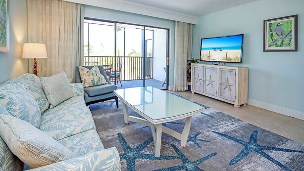 Ein Wohnzimmer mit Glastisch, einer Couch, einem Fernseher und einem Balkon mit Meerblick.
