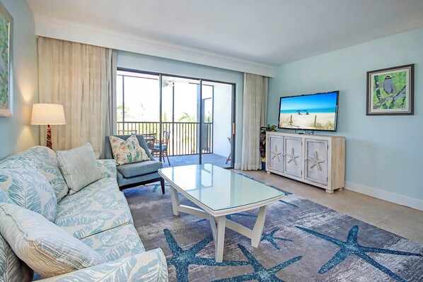 A living room with a glass-top coffee table, a sofa, a television, and a balcony with a view of the beach.