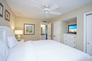 A hotel room with a large bed, a ceiling fan, a TV, and a view of the beach through an open door.