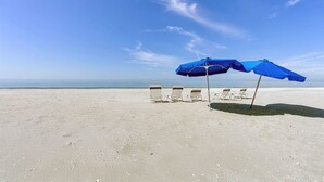 Ein Strand mit weißem Sand, drei Liegestühlen und zwei blauen Sonnenschirmen.