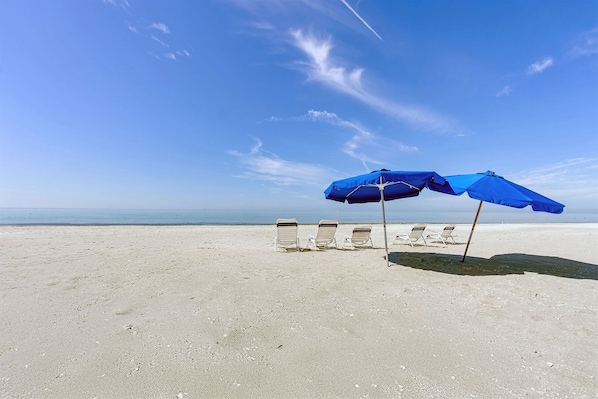 Une plage de sable blanc, avec trois chaises longues et deux parasols bleus.