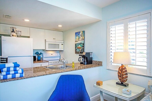 A modern kitchen with white cabinets, a microwave, and a coffee maker. There is a blue chair and a table with a decorative object.