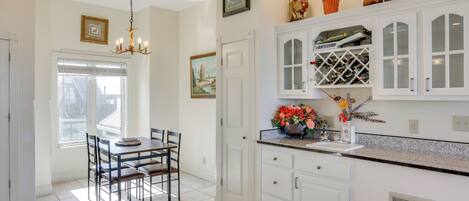 A kitchen with white cabinets, a dining table with chairs, a chandelier, and a window with blinds.