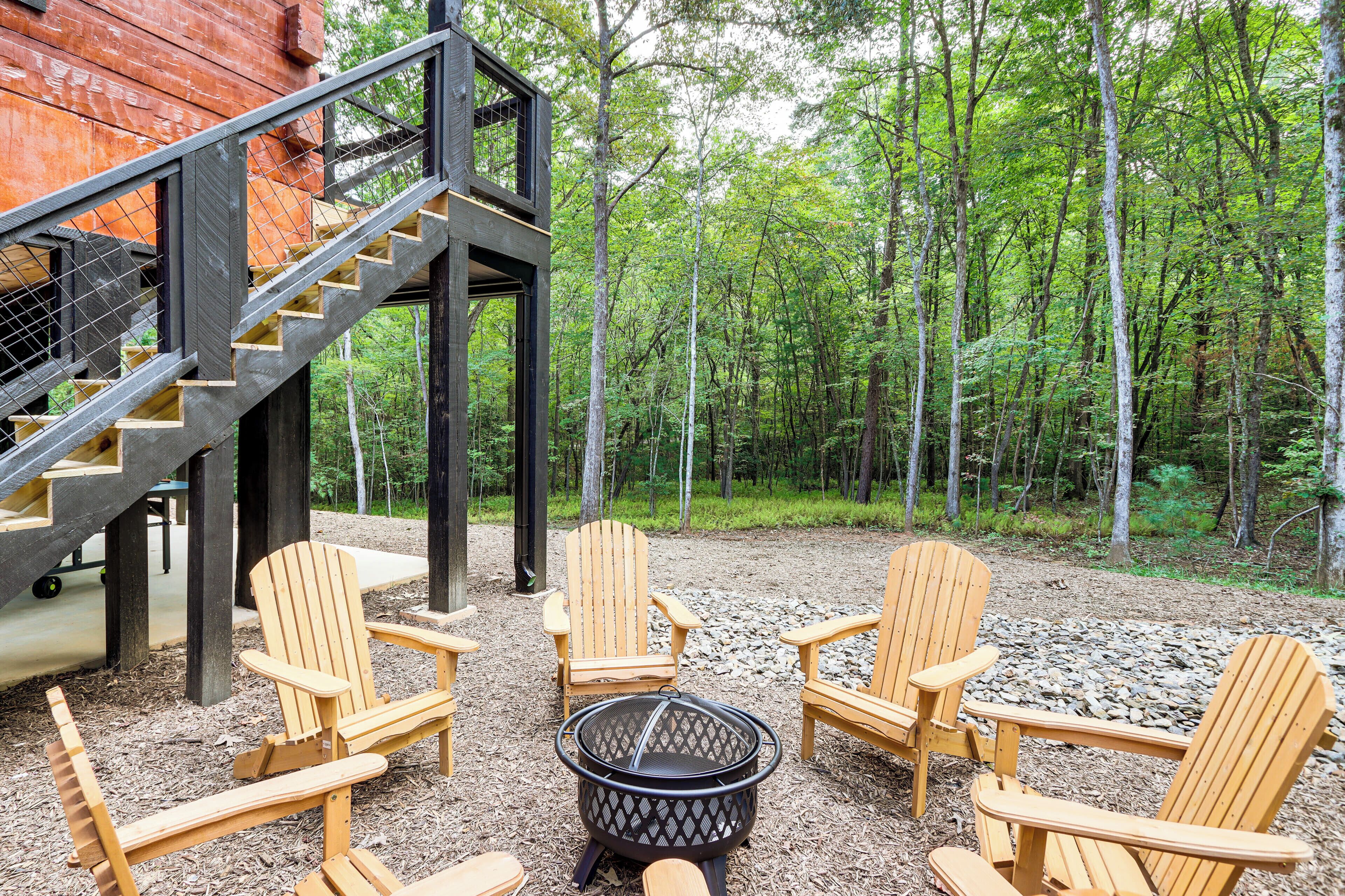 Een houten terras met Adirondack-stoelen en een vuurkuil, omringd door een bos.