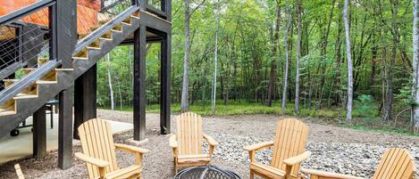 Een houten terras met Adirondack-stoelen en een vuurkuil, omringd door een bos.