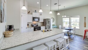 A modern kitchen with white cabinetry, granite countertops, and stainless steel appliances. There are pendant lights, a dining area with white chairs, and a large window providing natural light. - Home w/ Pool Access in Rockport! (Rockport)