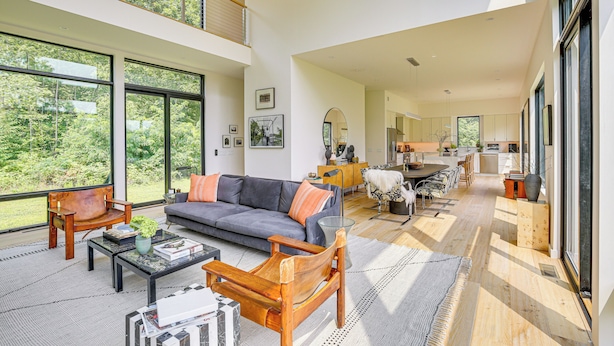 Een ruime woonkamer met een bank, salontafel en stoelen, grote ramen met uitzicht op groen, en een keuken op de achtergrond.