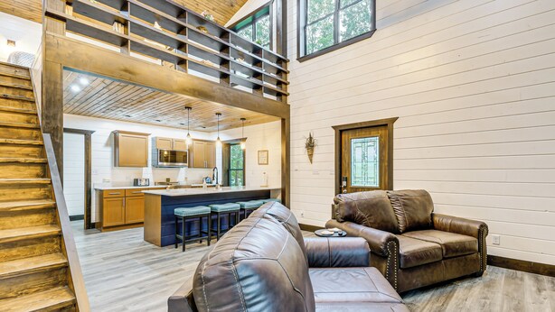 A spacious living area with leather sofas, a kitchen in the background, and a wooden staircase. - Red River Gorge Barndominium: Dog Friendly! (Beattyville)