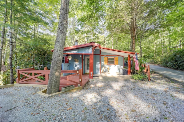A small, single-story cabin with a red and blue exterior, surrounded by trees and gravel.