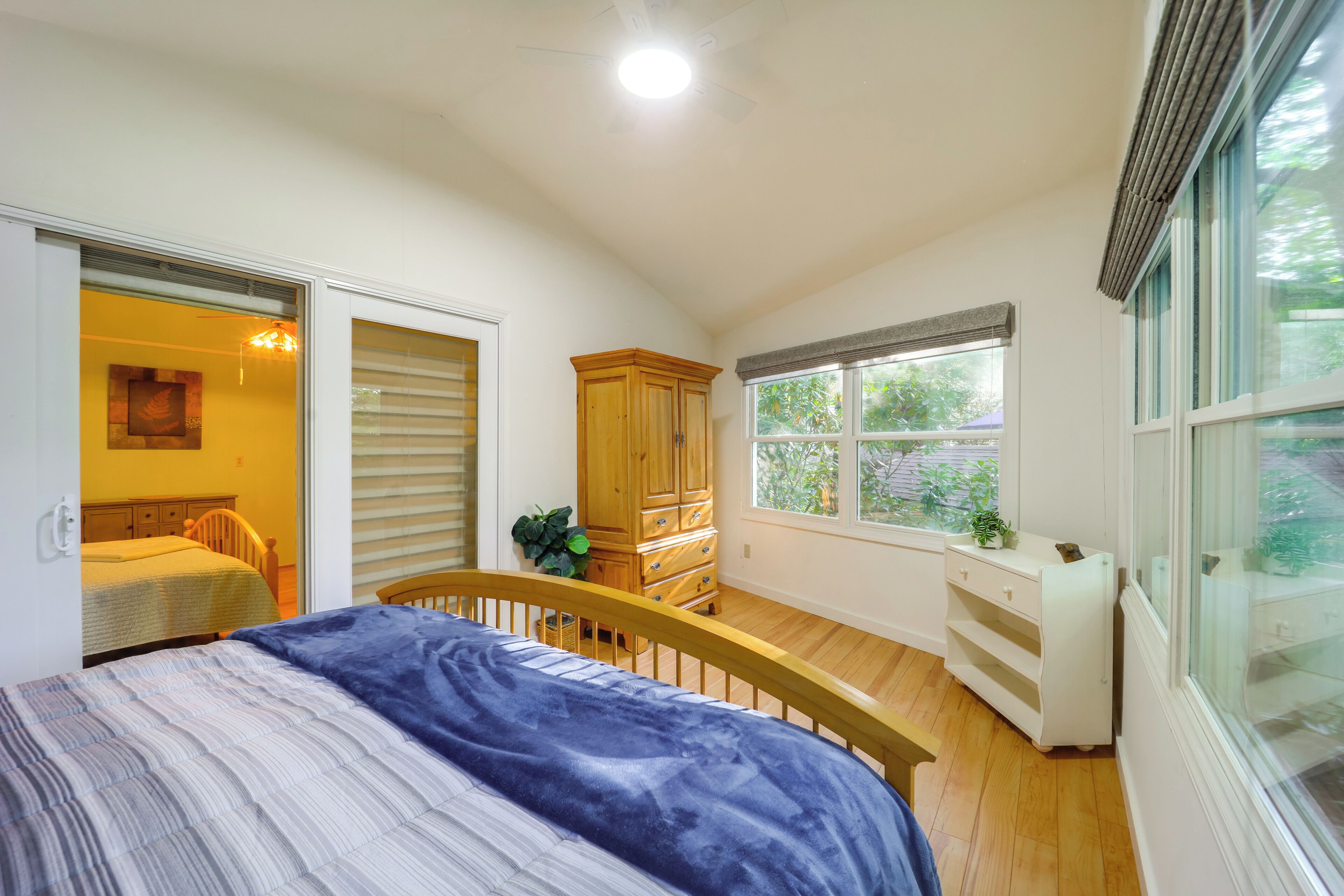 A bedroom with a bed, a wooden wardrobe, a window with a view of greenery, and a door leading to another room.