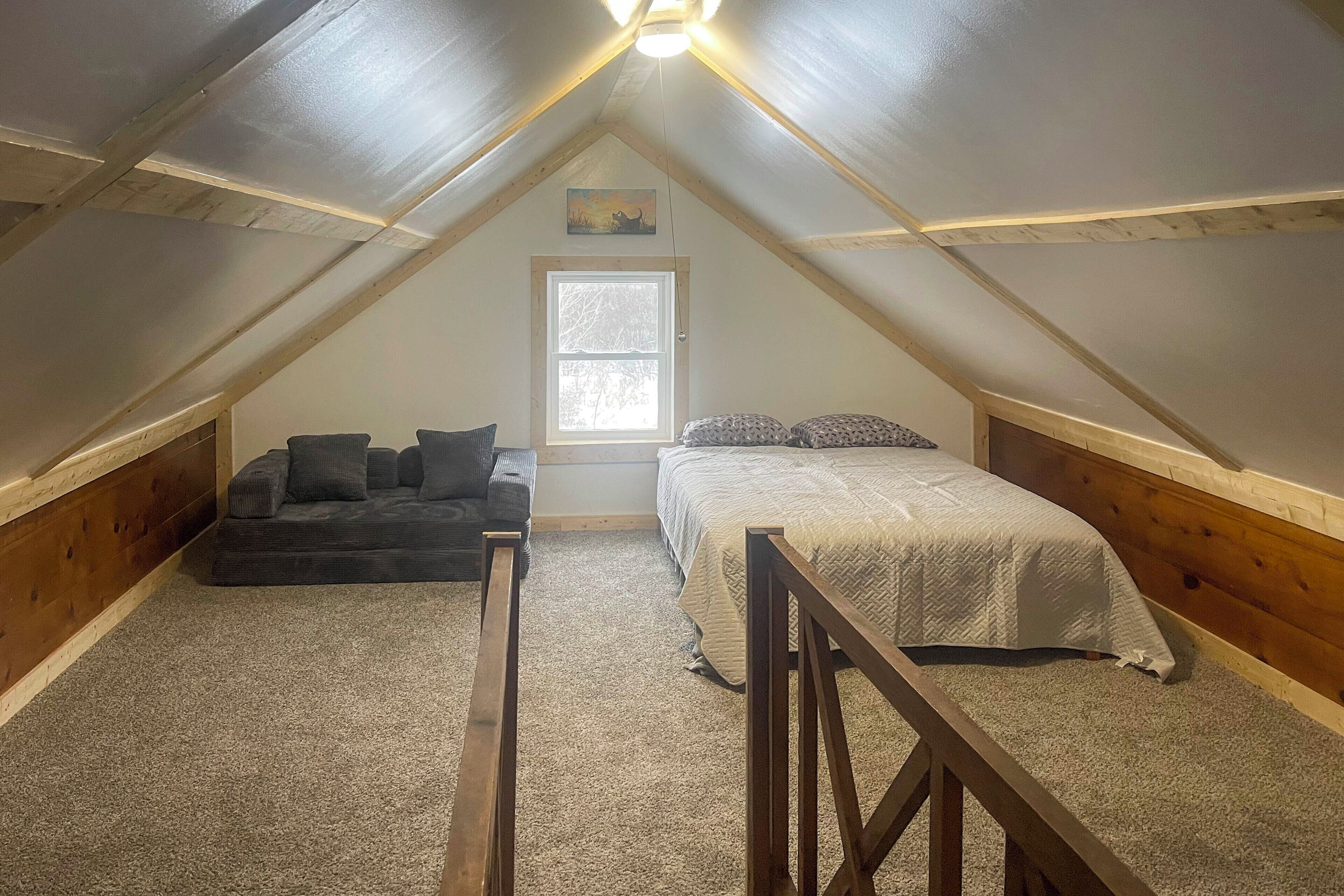 Un dormitorio en ático con techo inclinado, una ventana, una cama, un sofá y una barandilla de madera.
