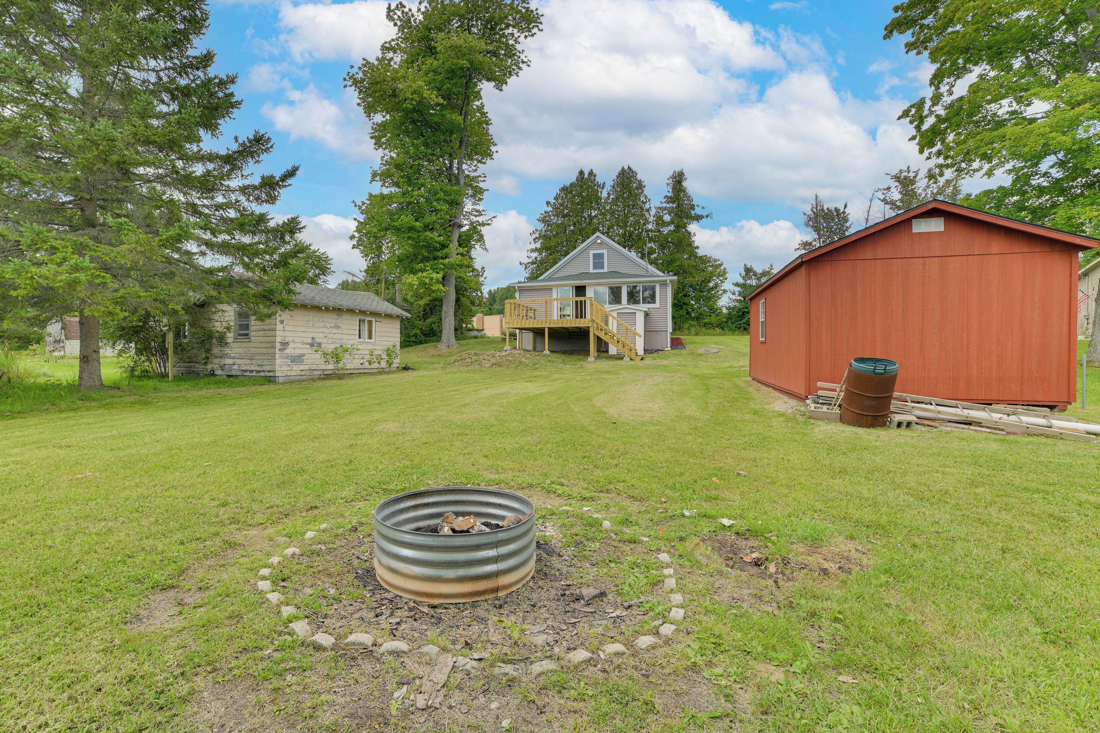 A rustic cabin with a wooden deck, a metal fire pit, and a red barn in a grassy yard with trees.