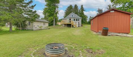 A rustic cabin with a wooden deck, a metal fire pit, and a red barn in a grassy yard with trees.