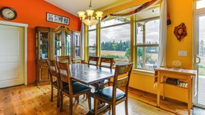 Uma sala de jantar com mesa e cadeiras de madeira, um lustre e vista para o exterior através de grandes janelas.