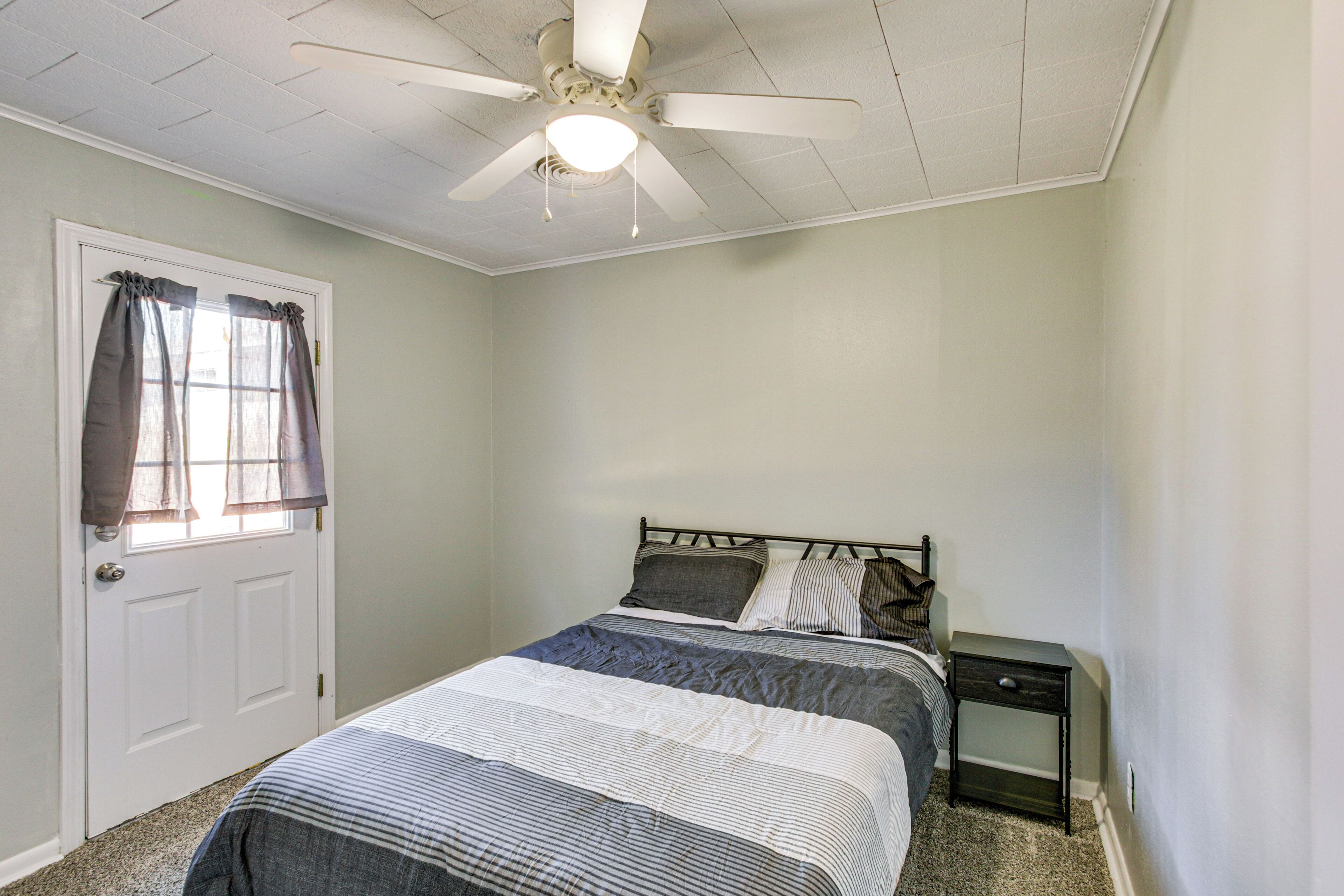 Un dormitorio con una cama, una mesita de noche, un ventilador de techo y una ventana con cortinas.