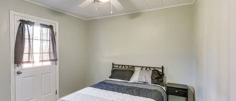 Un dormitorio con una cama, una mesita de noche, un ventilador de techo y una ventana con cortinas.