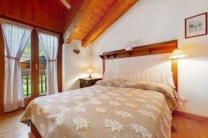 A bedroom with a wooden ceiling, a bed with a floral bedspread, two bedside lamps, and a window with curtains.