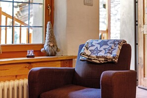 Ein Sessel mit Decke, ein Heizkörper, ein Holzregal, ein kleiner Weihnachtsbaum und ein Fenster mit Blick auf eine Holzhütte.