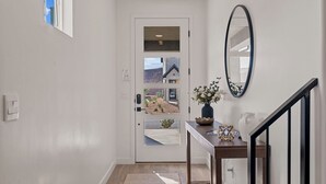 Un pasillo con una puerta blanca, un espejo, una mesita con un florero y una escalera de madera.