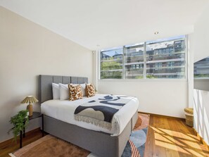 Ein modernes Schlafzimmer mit einem großen Bett, einem Nachttisch, einer Lampe, einem Fenster mit Blick auf Gebäude und einer kleinen Pflanze.
