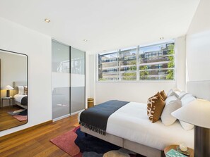 A modern bedroom with a large bed, a mirror, a nightstand, and a view of a building through the window.