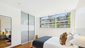 A modern bedroom with a large bed, a mirror, a nightstand, and a view of a building through the window. - Stunning Camden Town Balcony Apartment (Greater London)