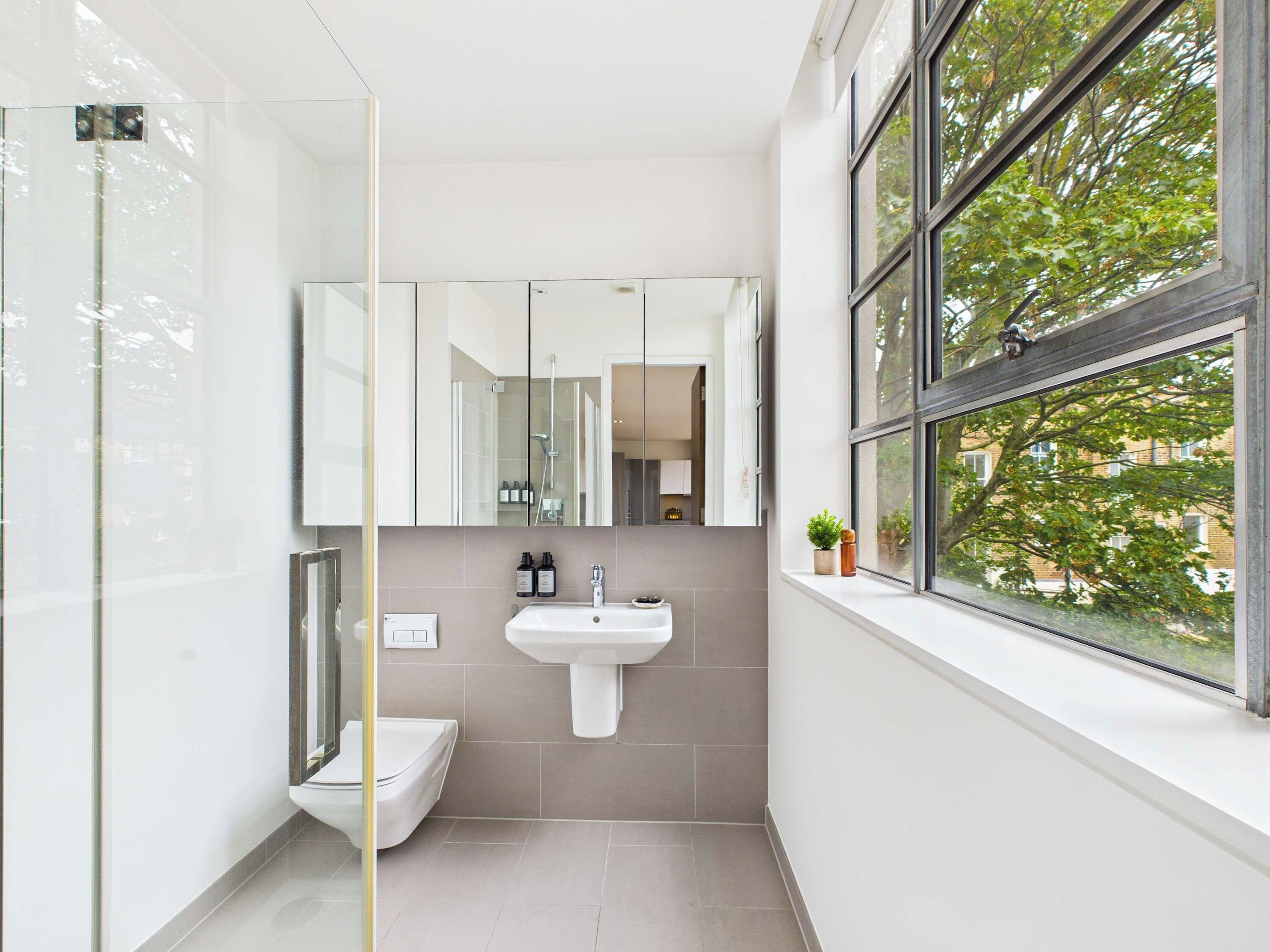 Een moderne badkamer met een glazen douchecabine, een wit wastafelmeubel en een raam met uitzicht op bomen.