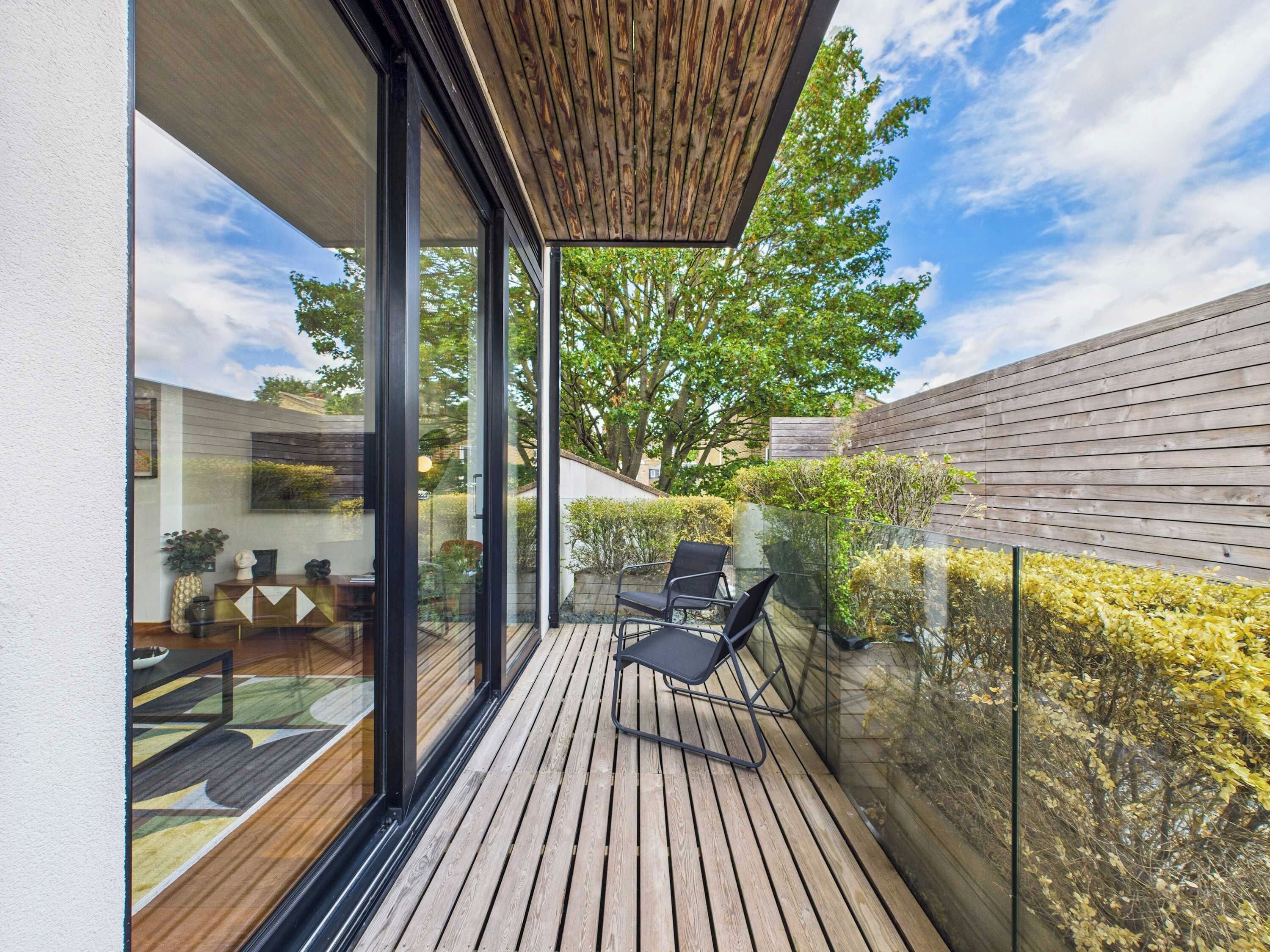 Un balcón moderno al aire libre con piso de madera, puertas de vidrio y vista a un árbol y edificios vecinos.