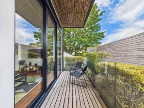 Un balcon extérieur moderne, doté d’un plancher en bois, de portes vitrées et offrant une vue sur un arbre et les bâtiments voisins.