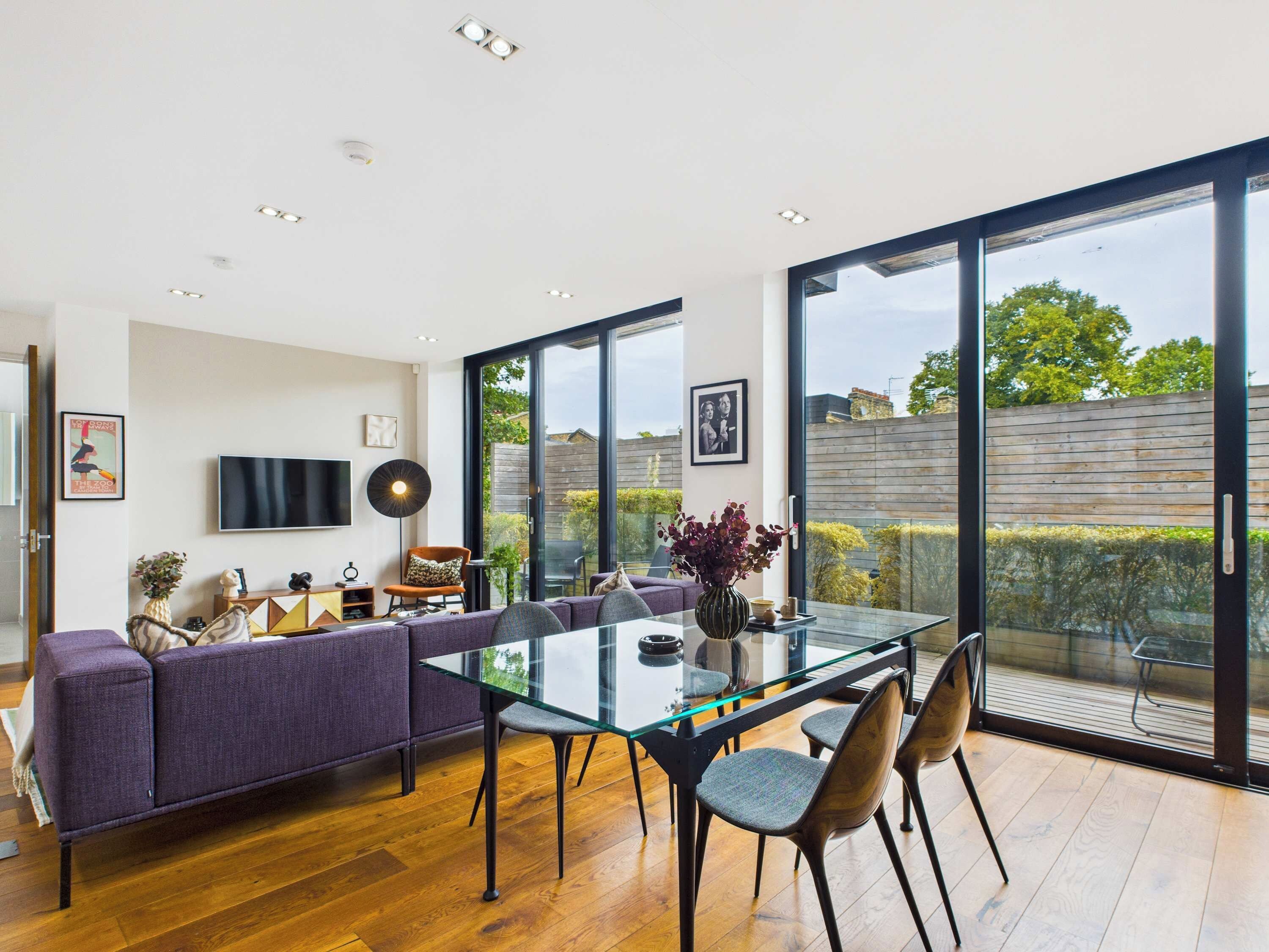 A modern dining area with a glass table, purple sofa, and a view of the garden through large windows.