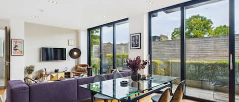 A modern dining area with a glass table, purple sofa, and a view of the garden through large windows.