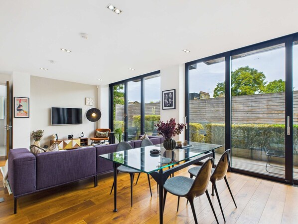A modern dining area with a glass table, purple sofa, and a view of the garden through large windows.