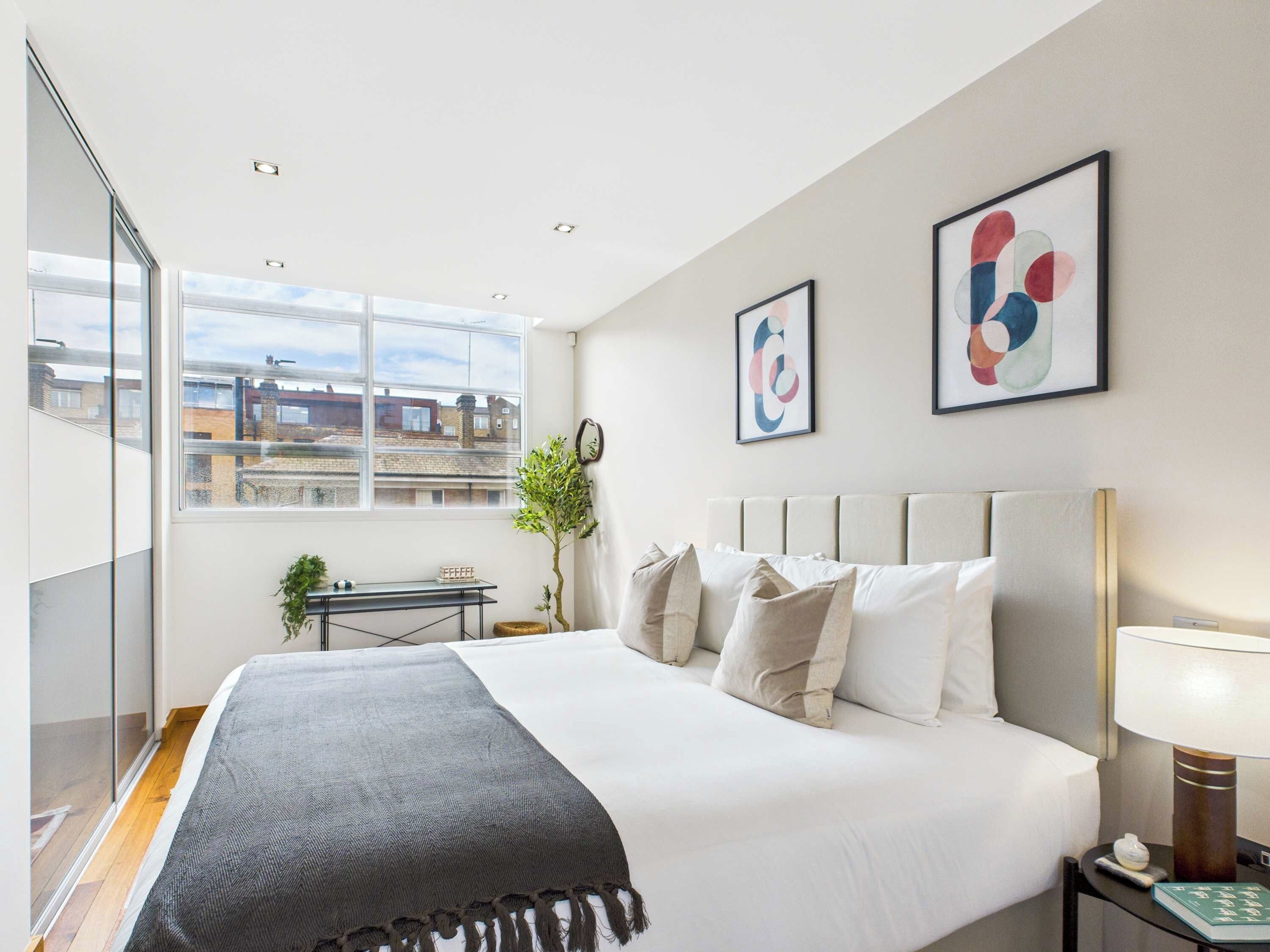 Une chambre d’hôtel moderne avec un grand lit, une table de chevet, une lampe et une fenêtre donnant sur des immeubles.