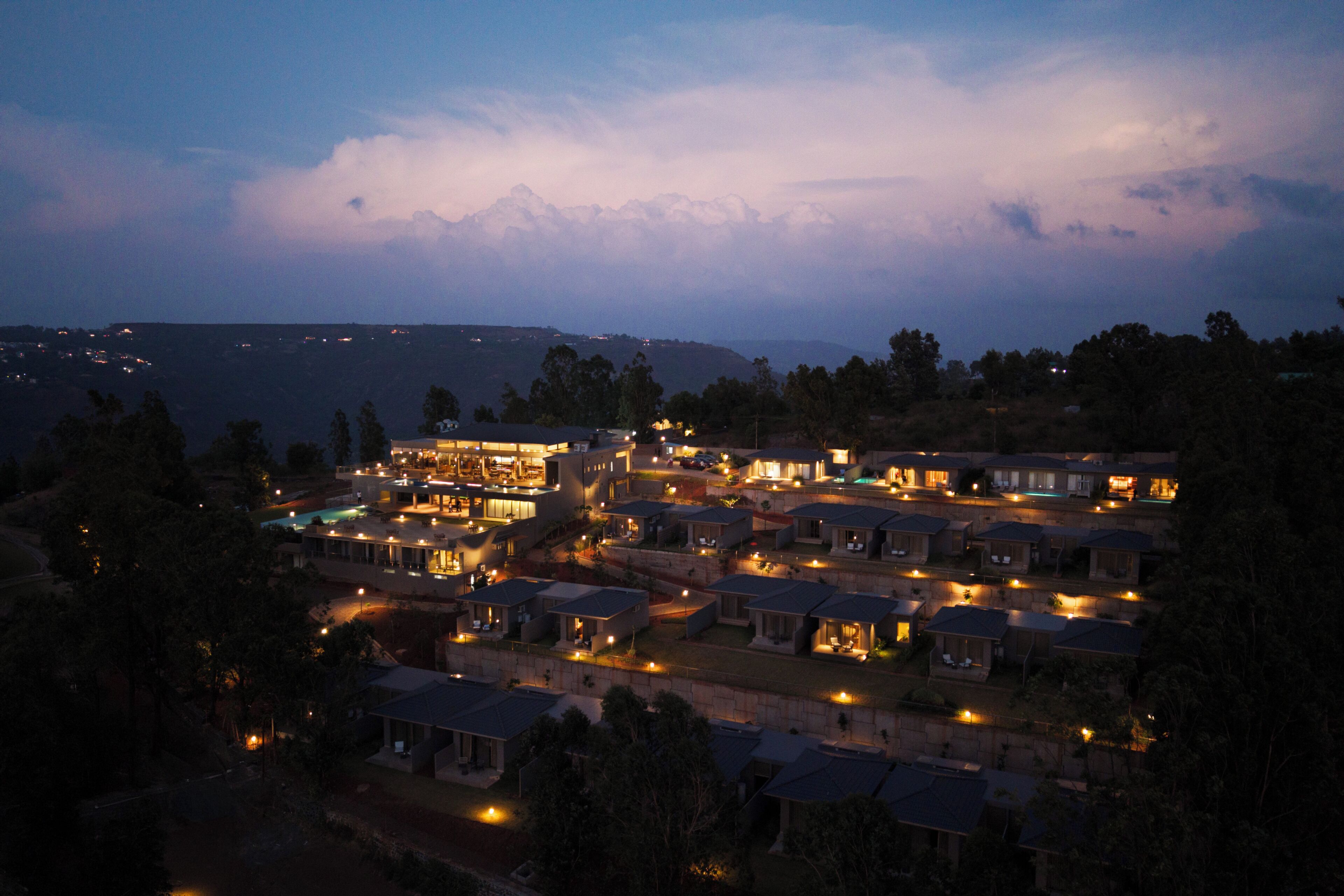 A residential area with multiple houses, some with visible outdoor lighting, surrounded by trees and hills in the background.