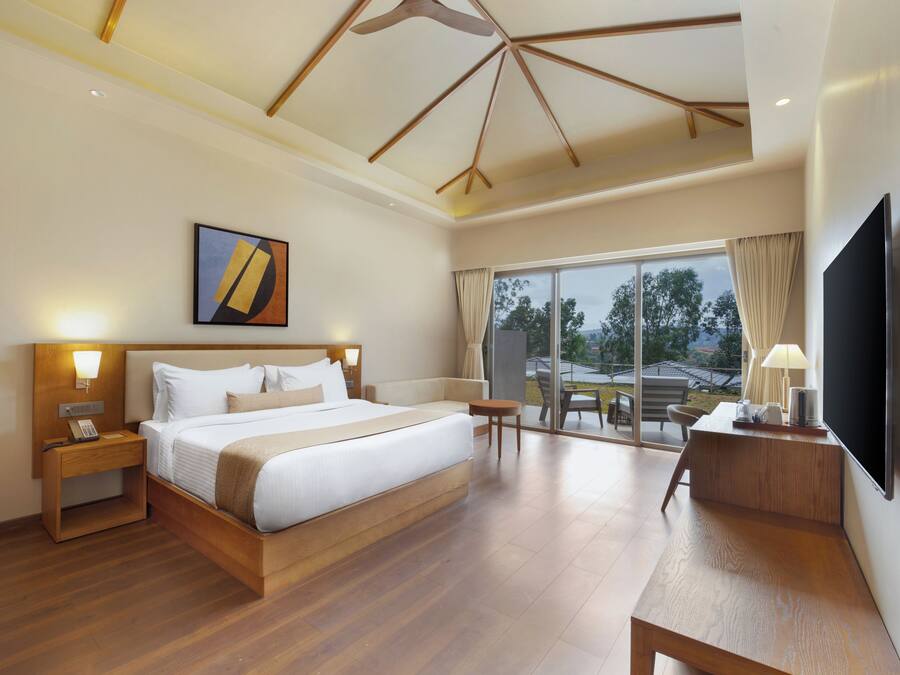 Habitación de hotel moderna con una cama grande, cabecero de madera, mesitas de noche con lámparas, un televisor de pantalla plana y un balcón con vistas al exterior.