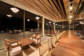 A spacious outdoor seating area with wooden tables and chairs, large windows, and hanging pendant lights. - Ayati Resort & Spa (Kaswand)