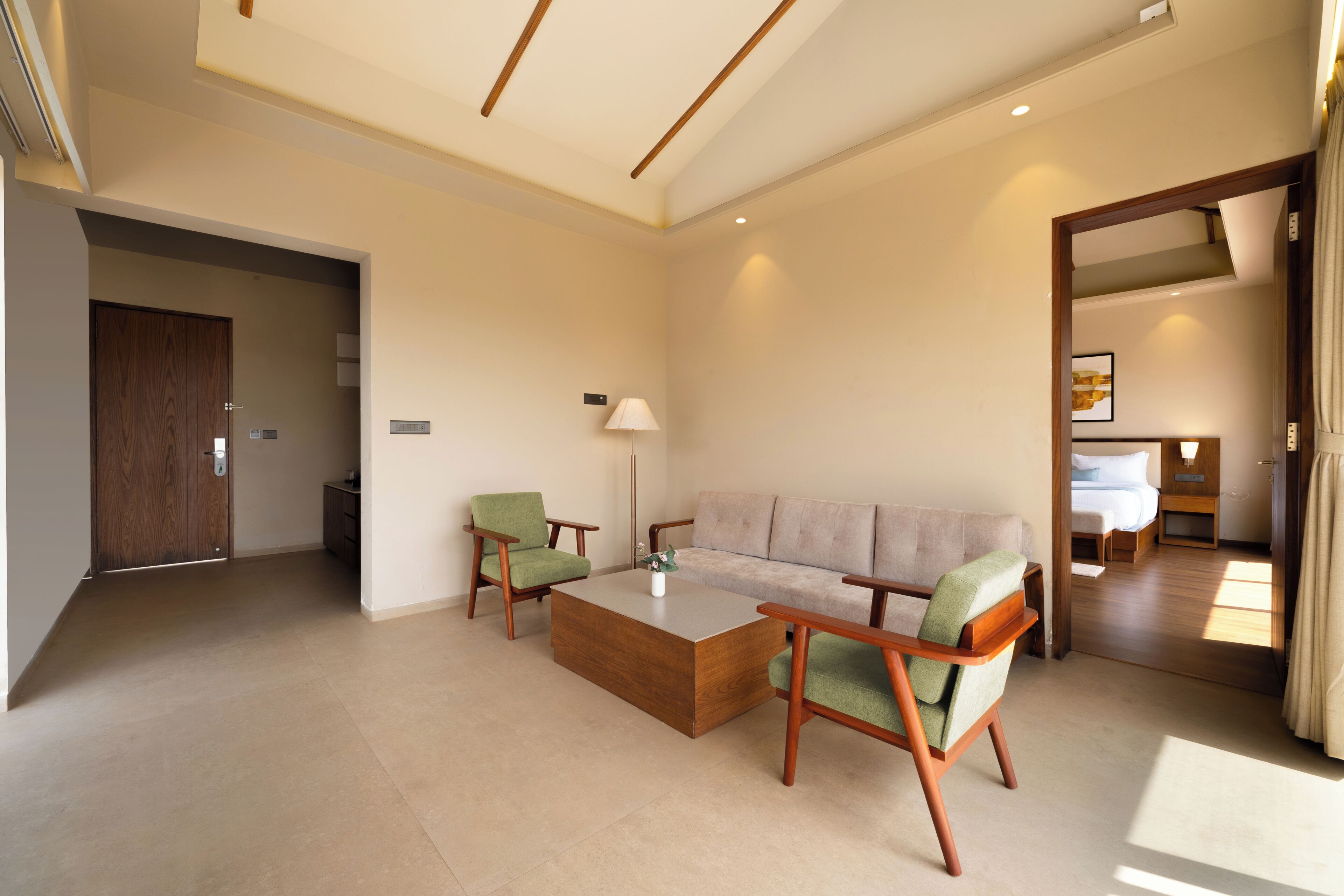 Une chambre d’hôtel moderne équipée d’un canapé, de deux fauteuils, d’une table basse, d’une lampe et offrant une vue sur la chambre adjacente.