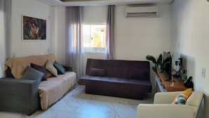 A living room with a sofa, a chair, and a coffee table. There is a painting on the wall and an air conditioning unit mounted on the wall.