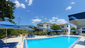 Une piscine entourée de bâtiments dotés de balcons, avec des chaises longues bleues et des parasols, le tout sur fond de paysage montagneux.