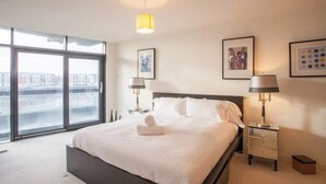 Une chambre d’hôtel moderne avec un grand lit, deux tables de chevet avec des lampes, une fenêtre donnant sur la ville et des œuvres d’art encadrées accrochées aux murs.
