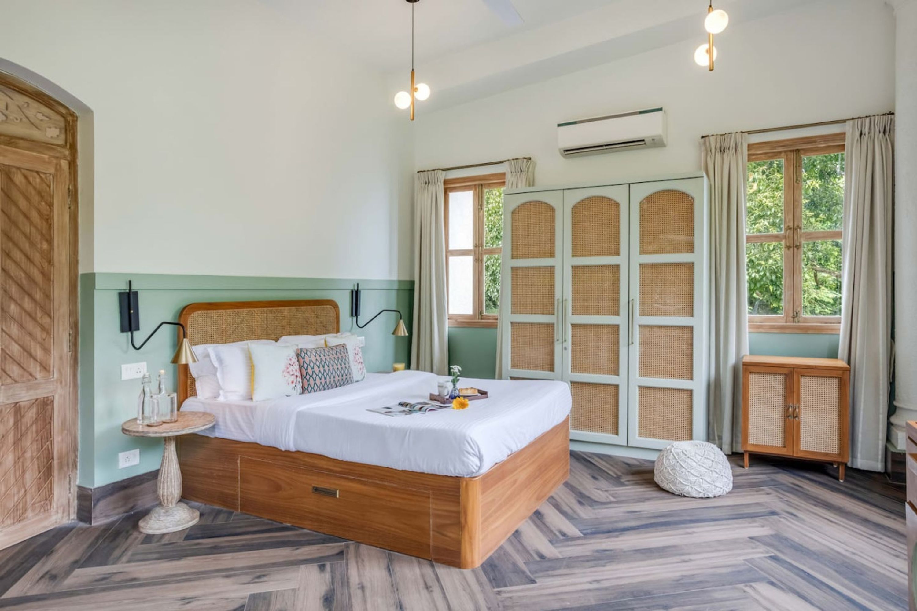 A bedroom with a wooden bed, a round nightstand, a wicker cabinet, and a large window with curtains.