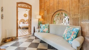 Een kamer met een geruite vloer, een lichtkleurige bank met bloemkussens, een houten wand met een rond raam, en een bed met een gestreept hoofdeinde op de achtergrond.