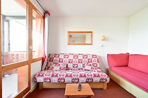 A room with a sofa bed, a small table with a remote control, a mirror, and a lamp. - Apartment Rental in La Plagne - Montchavin - Les Coches (Bellentre)