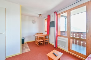 A room with a wooden table and chairs, a small wooden table with a remote, a window with a view, and a door.